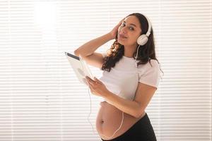 hermosa joven embarazada elige música por suscripción y la escucha a través de auriculares y tabletas por la mañana. concepto de beneficios para el desarrollo fetal foto