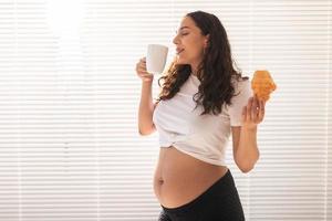 Pregnant woman eating croissant and drinks coffee. Pregnancy and maternity leave. Copy space photo