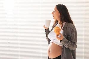 Healthy beautiful pregnant woman drinking tea and eating croissant during lunch. Concept of high-calorie nutrition while waiting for birth of baby. Copy space. photo