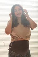 Curly-haired brunette pacified pregnant woman listens to pleasant classical music using smartphone and headphones. Concept of a soothing mood before meeting baby. photo