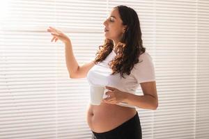 Curious pregnant woman drinking tea and looking through the blinds at the window. Concept of joy and good news while waiting for baby photo