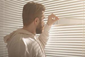 Rear view of young man with beard peeks through hole in the window blinds and looks out into the street. Surveillance and curiosity concept photo