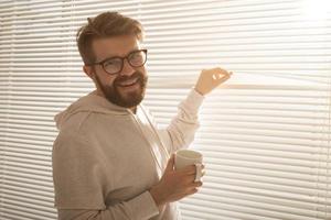 Young stylish man with beard and coffee peeking through hole in window blinds and looking out into street. Concept of enjoying the morning sun and positivity photo