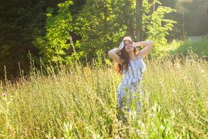 mujer joven feliz con el pelo largo con sombrero y vestido caminando por el bosque de verano en un día soleado. concepto de alegría de verano foto