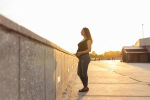 Portrait of hispanic pregnant woman walking on embankment at sunset. Pregnancy and motherhood concept. Advertising area workspace photo