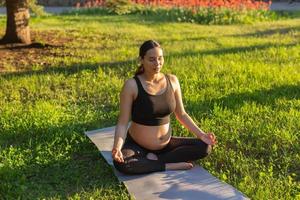 Peaceful young positive pregnant woman in gymnastic suit does yoga and meditate sitting on mat on green grass on sunny warm summer day. Concept of preparation for childbirth and positive attitude photo