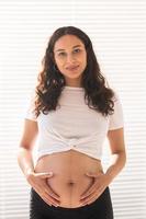 Smiling young beautiful pregnant woman touching her belly and rejoicing. Concept of positive and pleasant feelings while waiting for the baby photo