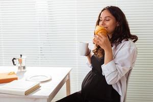 Happy pregnant young beautiful woman eating croissant during morning breakfast. Concept of pleasant morning and positive attitude during pregnancy photo