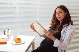 Side view of happy young beautiful woman reading book while having morning breakfast with coffee and croissants on background of blinds. Good morning concept and pleasant lunch break photo