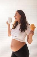 Happy pregnant young beautiful woman eating croissant during morning breakfast. Concept of pleasant morning and positive attitude during pregnancy photo