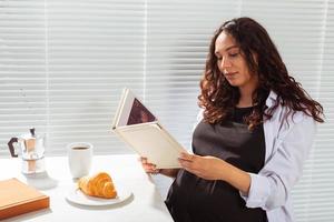 Side view of pregnant woman reading book while having morning breakfast with coffee and croissants on background of blinds. Good morning concept and pleasant lunch break photo