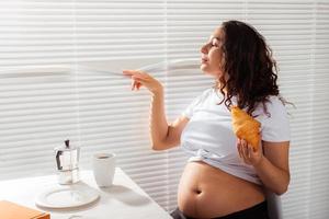 feliz embarazada joven hermosa mujer comiendo croissant y mirando a través de las persianas durante el desayuno de la mañana. concepto de mañana agradable y actitud positiva durante el embarazo foto