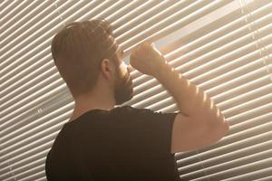 la vista trasera de un joven con barba se asoma por un agujero en las persianas de la ventana y mira hacia la calle. concepto de vigilancia y curiosidad foto