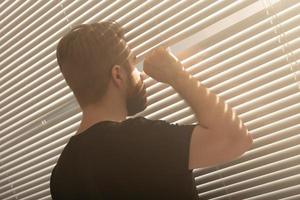 Rear view of young man with beard peeks through hole in the window blinds and looks out into the street. Surveillance and curiosity concept photo