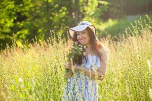 Happy young woman with long hair in hat and dress pulls her hands towards the plants while walking through the summer forest on a sunny day. Summer joy concept photo