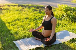 Una joven y pacífica mujer embarazada positiva con traje de gimnasia hace yoga y medita sentada en una alfombra sobre hierba verde en un día soleado y cálido de verano. concepto de preparación para el parto y actitud positiva foto