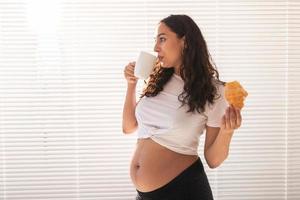 Happy pregnant young beautiful woman eating croissant during morning breakfast. Concept of pleasant morning and positive attitude during pregnancy. Copy space. photo