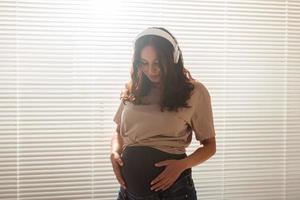 morena sonriente mujer embarazada pacificada escuchando música agradable usando un teléfono inteligente y auriculares. concepto de terapia de sedación antes de reunirse con el bebé foto