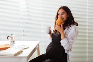 Happy pregnant young beautiful woman eating croissant during morning breakfast. Concept of pleasant morning and positive attitude during pregnancy photo