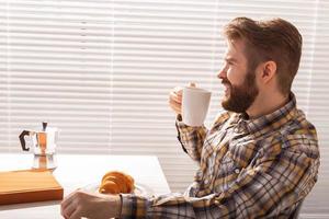 Side view of pensive young bearded male businessman drinking cup of coffee on background of blinds. Concept of pleasant morning or lunch break. Copyspace photo