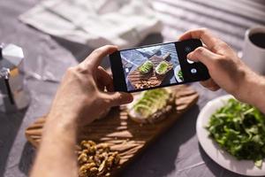 Hands take pictures on smartphone of two beautiful healthy sour cream and avocado sandwiches lying on board on the table. Social media and food concept photo