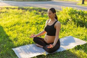 Peaceful young positive pregnant woman in gymnastic suit does yoga and meditate sitting on mat on green grass on sunny warm summer day. Concept of preparation for childbirth and positive attitude photo
