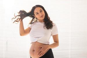 Smiling young beautiful pregnant woman touching her belly and hair and rejoicing. Concept of health and thinking about the future while waiting for baby. Copyspace photo