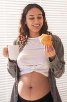 hermosa mujer embarazada sana bebiendo té y comiendo croissant durante el almuerzo. concepto de nutrición alta en calorías mientras se espera el nacimiento del bebé foto