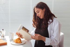 Side view of happy young beautiful woman reading book while having morning breakfast with coffee and croissants on background of blinds. Good morning concept and pleasant lunch break photo