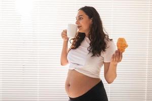 Beautiful pregnant woman holding croissant and cup of coffee in her hands during morning breakfast. Concept of good health and positive attitude while expecting baby. Copy space. photo