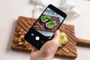 Hands take pictures on smartphone of two beautiful healthy sour cream and avocado sandwiches lying on board on the table. Social media and food concept photo