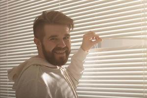 Young man with beard peeks through hole in the window blinds and looks out into the street. Surveillance and curiosity concept photo