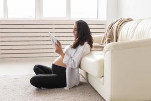 Pregnant woman holding tablet sitting on a carpet near a couch in the living room at home photo