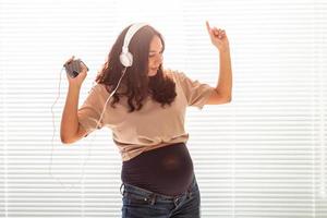 Curly-haired brunette pacified pregnant woman listens to pleasant classical music using smartphone and headphones. Concept of a soothing mood before meeting baby.