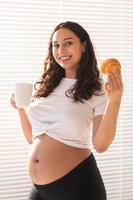 hermosa mujer embarazada sosteniendo un croissant y una taza de café en sus manos durante el desayuno de la mañana. concepto de buena salud y actitud positiva mientras se espera un bebé foto