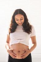 Smiling young beautiful pregnant woman touching her belly and rejoicing. Concept of positive and pleasant feelings while waiting for the baby photo