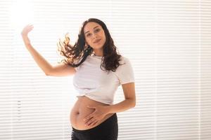 Smiling young beautiful pregnant woman touching her belly and hair and rejoicing. Concept of health and thinking about the future while waiting for baby. Copyspace photo