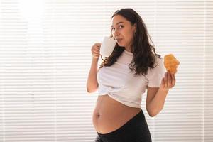 Pregnant woman eating croissant and drinks coffee. Pregnancy and maternity leave. Copy space photo