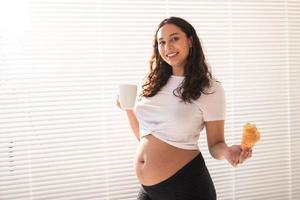 hermosa mujer embarazada sosteniendo un croissant y una taza de café en sus manos durante el desayuno de la mañana. concepto de buena salud y actitud positiva mientras se espera un bebé foto