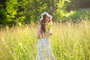 Happy young woman with long hair in hat and dress pulls her hands towards the plants while walking through the summer forest on a sunny day. Summer joy concept photo