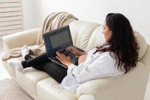 Latin pregnant woman using laptop computer sitting on sofa at home. Pregnancy and information for parenthood concept. photo