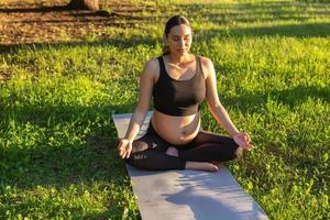 Peaceful young positive pregnant woman in gymnastic suit does yoga and meditate sitting on mat on green grass on sunny warm summer day. Concept of preparation for childbirth and positive attitude photo