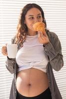 hermosa mujer embarazada sana bebiendo té y comiendo croissant durante el almuerzo. concepto de nutrición alta en calorías mientras se espera el nacimiento del bebé foto