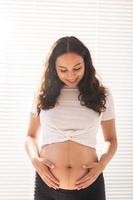 Smiling young beautiful pregnant woman touching her belly and rejoicing. Concept of positive and pleasant feelings while waiting for the baby photo