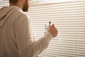 Rear view of young stylish man peeking through hole in window blinds and looking out into street. Concept of enjoying the morning sun and positivity photo