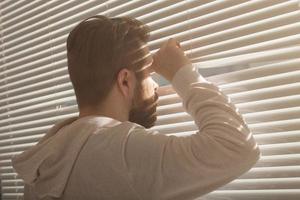 Rear view of young man with beard peeks through hole in the window blinds and looks out into the street. Surveillance and curiosity concept photo
