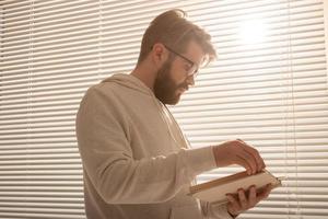 Bottom view of pensive stylish hipster man leafing through his daily planner in front of the blinds. Planning and time saving concept photo