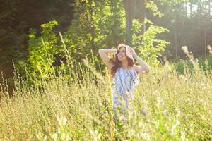 mujer joven feliz con el pelo largo con sombrero y vestido caminando por el bosque de verano en un día soleado. concepto de alegría de verano foto