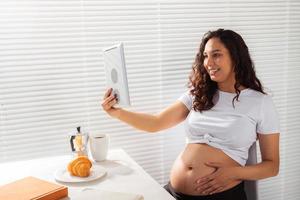 Happy pregnant young beautiful woman talking to mom using video call during morning breakfast. Concept of communication and positive attitude during pregnancy photo