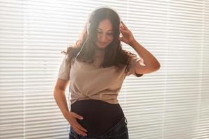 Portrait of pregnant woman near window at home and listening music in headphones. Pregnancy and leisure concept. photo
